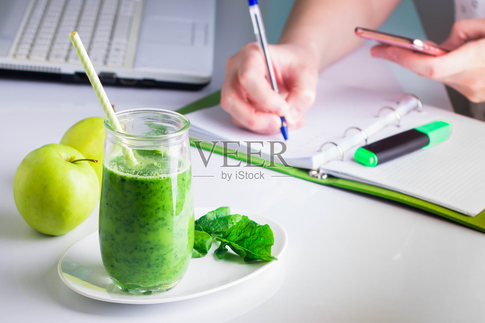 女人右手在笔记本上写字，左手拿着手机手机智能手机，笔记本电脑在旁边。来自绿色菠菜和苹果的健康饮料奶昔照片摄影图片