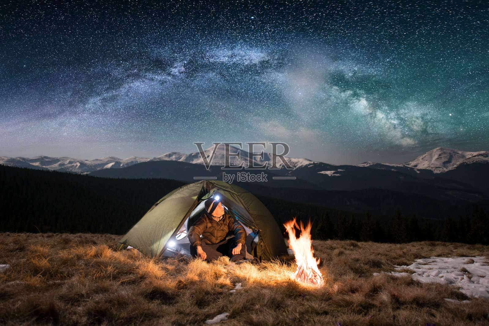 男游客晚上在山中露营休息。在满是星星和银河的美丽夜空下，一个带着头灯的男人坐在篝火和帐篷旁，欣赏着夜景照片摄影图片