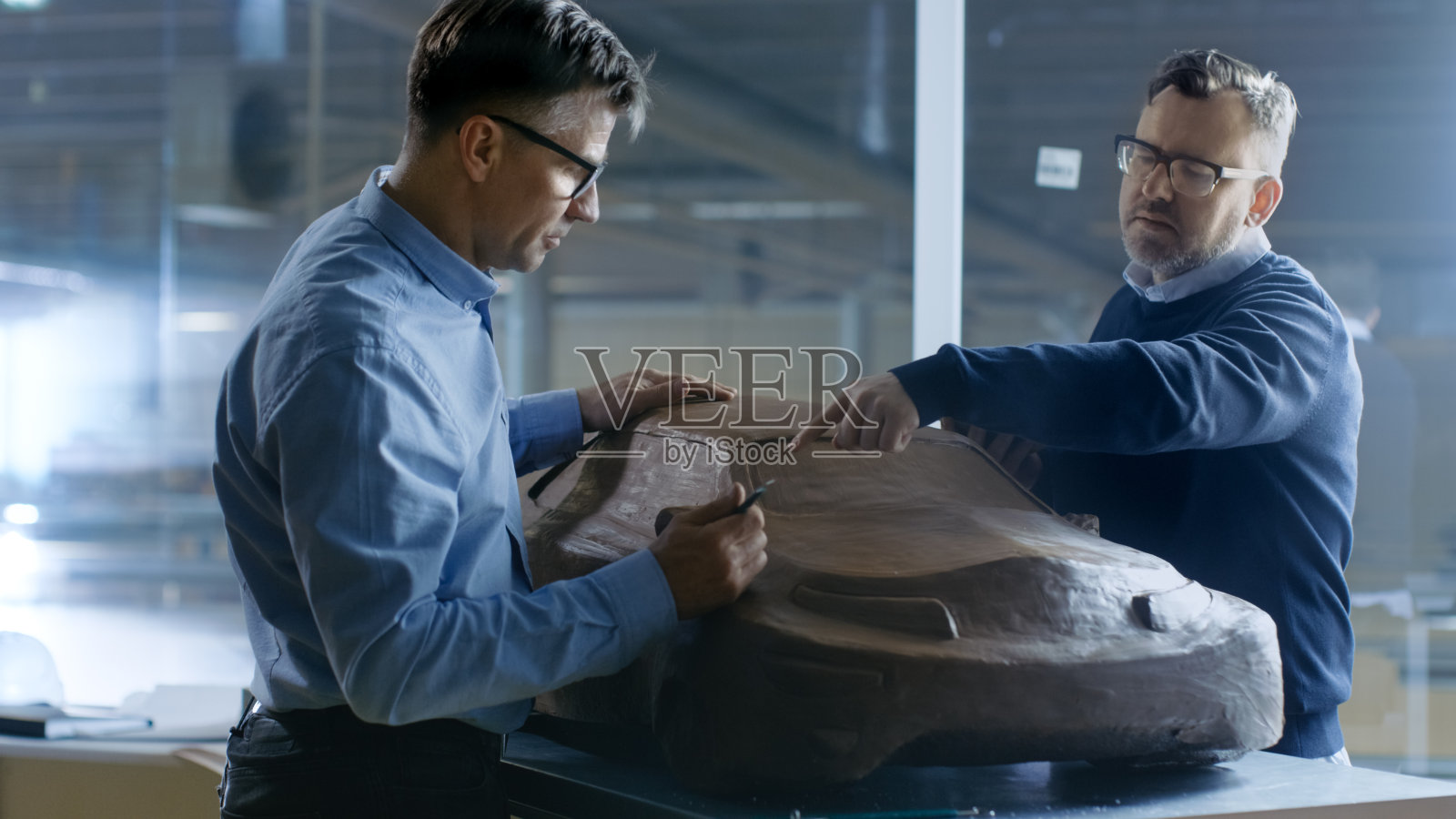 两位男性汽车设计师正在研究新一代电动汽车未来设计的泥塑模型。一个持有平板电脑图形设计，其他雕塑与粘土与耙子/线。照片摄影图片