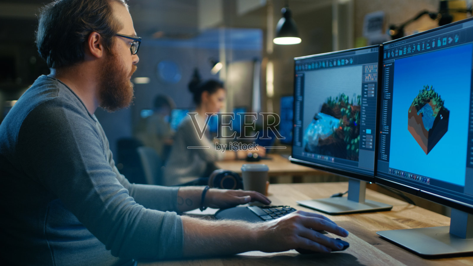富有创造性的男性游戏开发者致力于新关卡的图像设计，他坐在自己的木桌上在两台显示个人电脑上工作。其他人在这个创意工作室工作。照片摄影图片