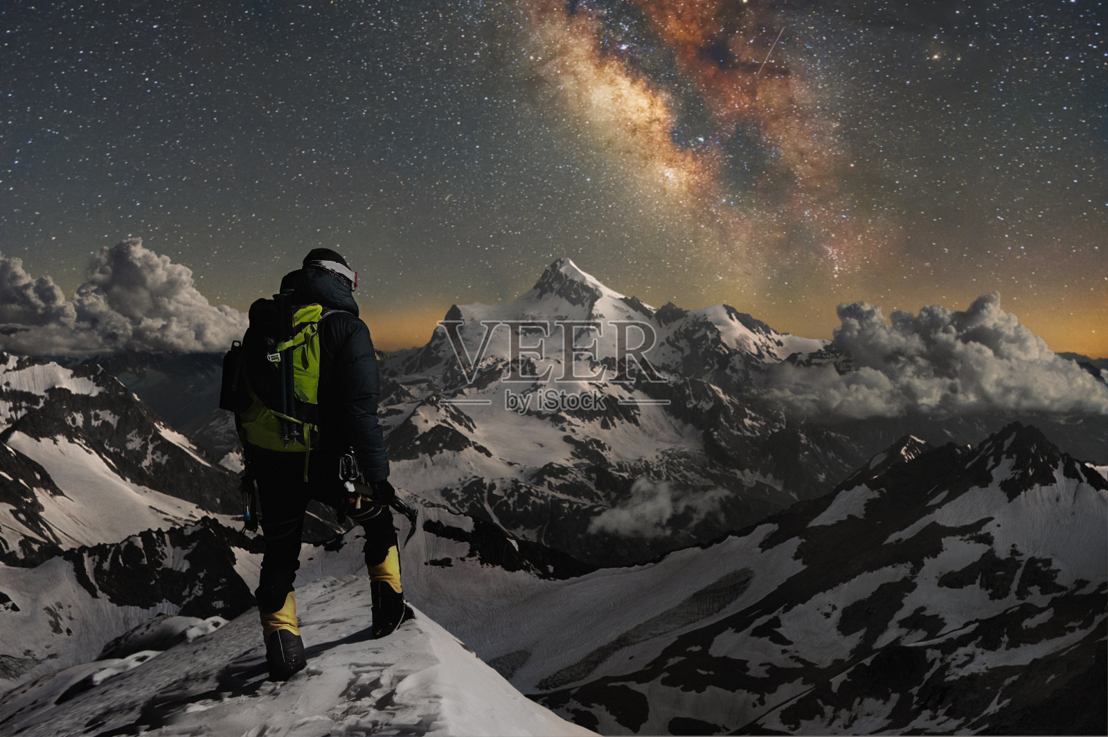 夜晚的照片登山者站在山顶的雪山，看着周围的山，星空和银河照片摄影图片