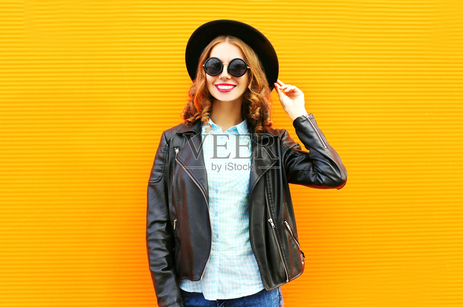 时尚微笑的女人在黑色岩石夹克，帽子在鲜艳的橙色背景照片摄影图片