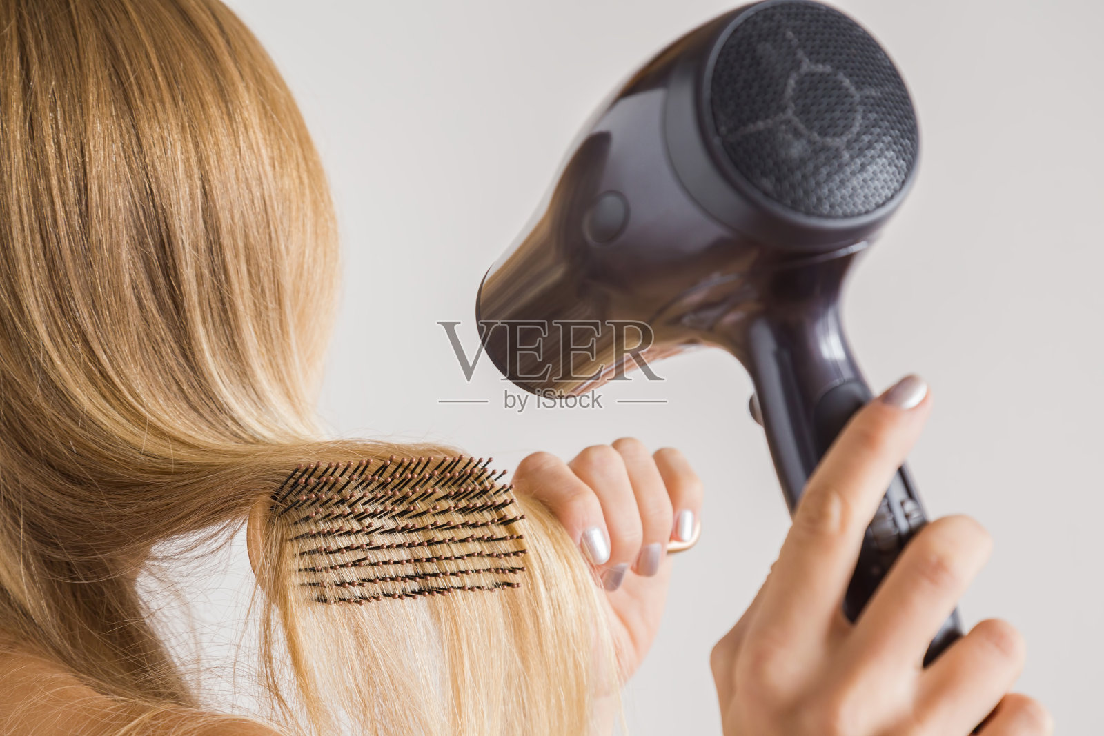 一个女人正在用吹风机和梳子梳理她灰色背景上的金发。关心健康清洁的头发。美容院的概念。照片摄影图片