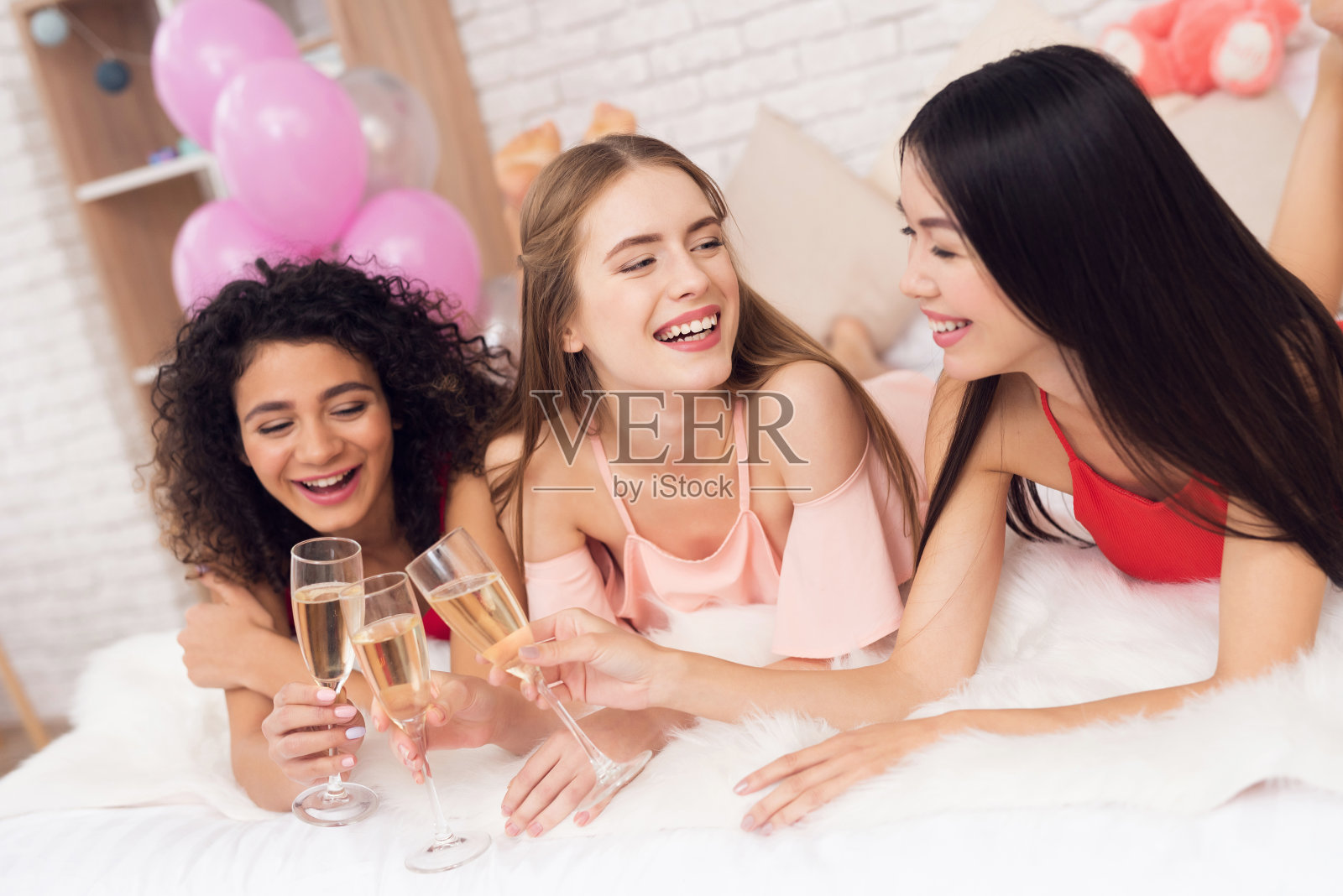 三个女孩躺在床上喝着香槟。他们正在庆祝3月8日的妇女节。照片摄影图片