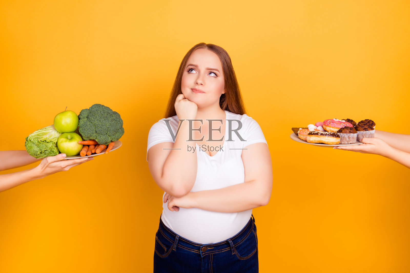 身体护理概念。困惑、深思、犹豫的胖女人无法在健康的天然产品和甜可口的饼干之间做出选择，孤立在明亮的黄色背景照片摄影图片