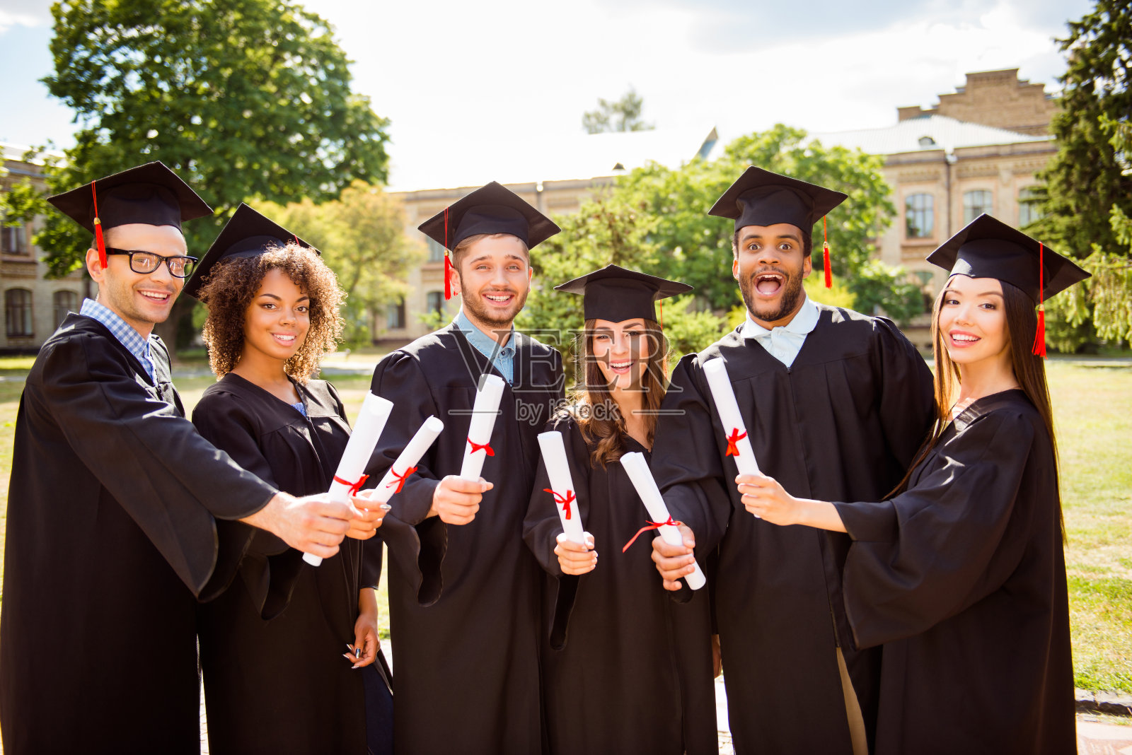 六名成功迷人的年轻毕业生身着黑色长袍，戴着帽子完成了他们的学业，手里拿着毕业证书，面带微笑，身后是学校大楼，阳光明媚照片摄影图片