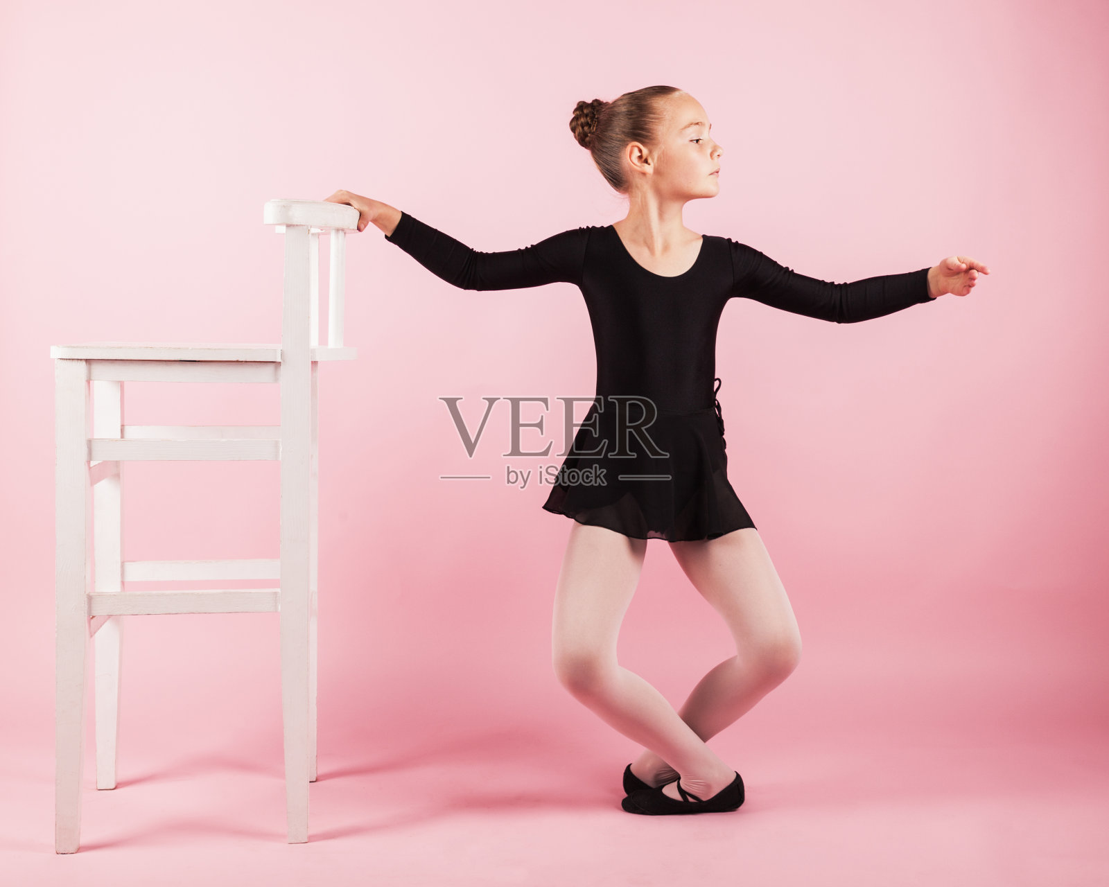 年轻美丽的女孩芭蕾舞女演员站在扶手椅附近练习芭蕾舞穿着黑色芭蕾舞裙摆在工作室与浅粉色的背景。照片摄影图片