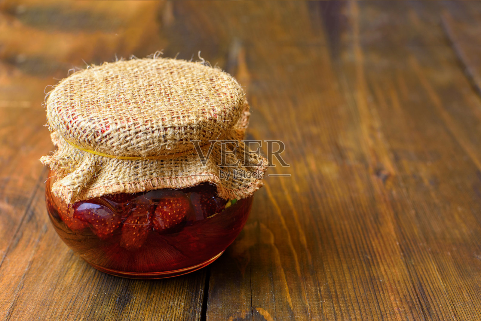 草莓果酱罐，盖子是用粗麻布做的，深色的木头做的背景照片摄影图片