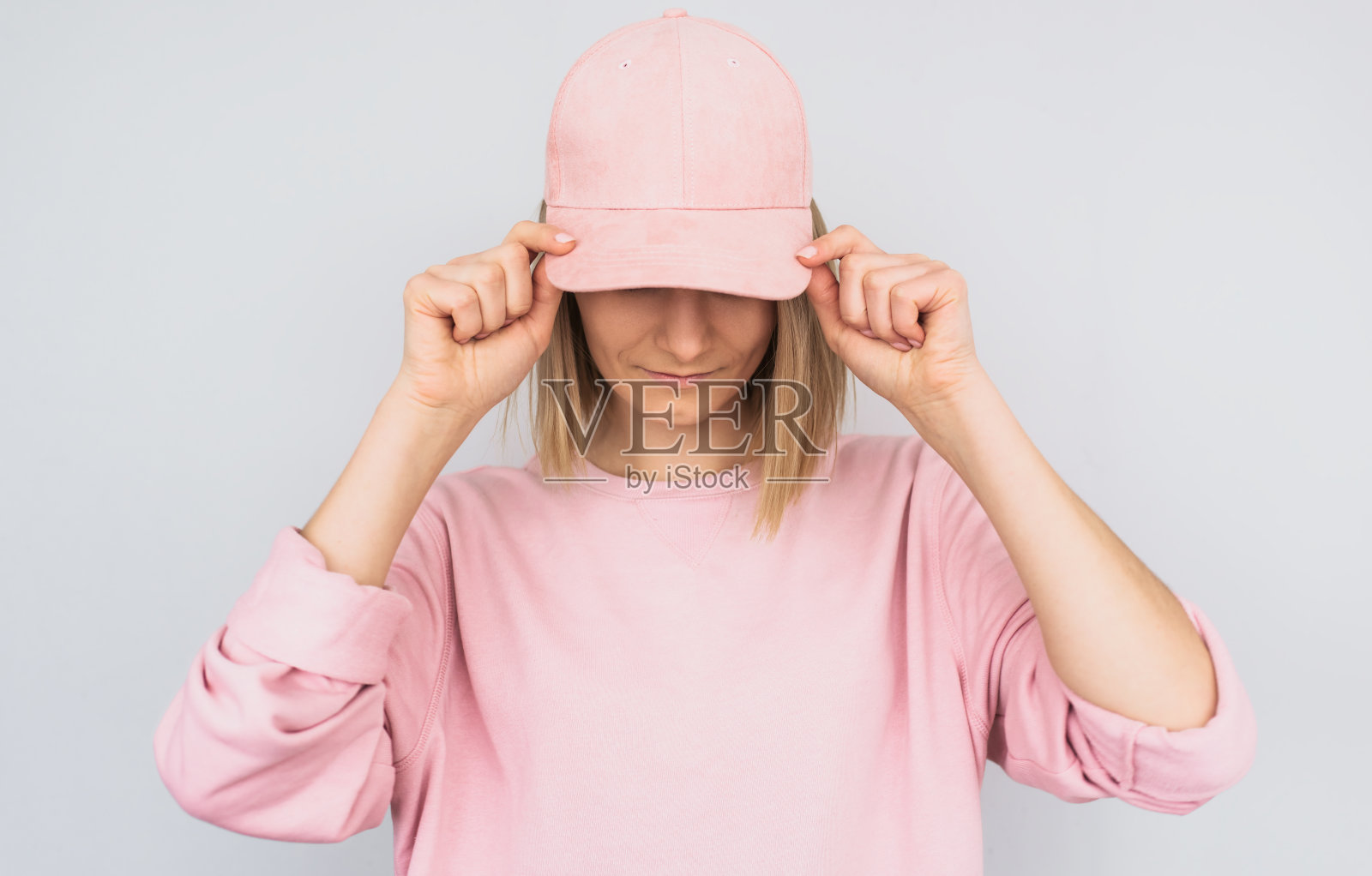 特写的白人金发年轻女性穿着粉红色的毛衣，用粉红色的帽子遮住她的眼睛，防止阳光，隔离在白色的工作室背景。积极的人的情感概念照片摄影图片