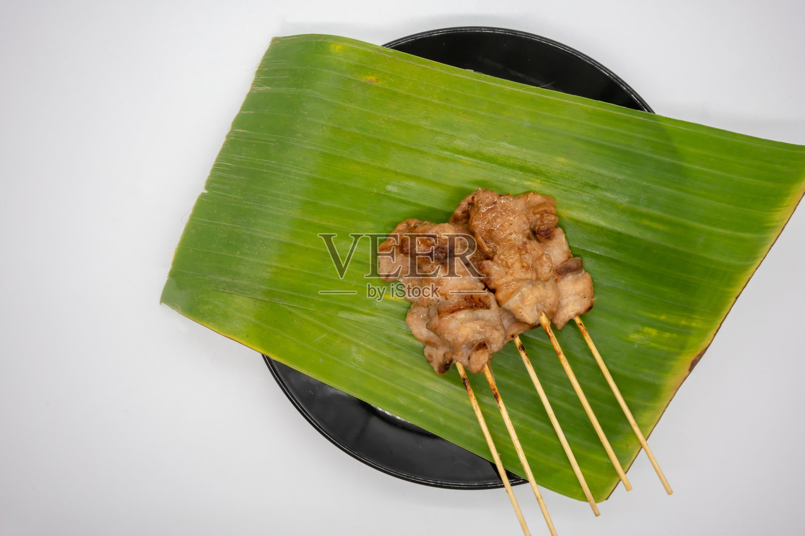 泰式烧烤或“Moo Ping”在泰国的单词。香蕉叶上用木棒塞的腌猪肉，白色背景上的黑色陶瓷盘。照片摄影图片