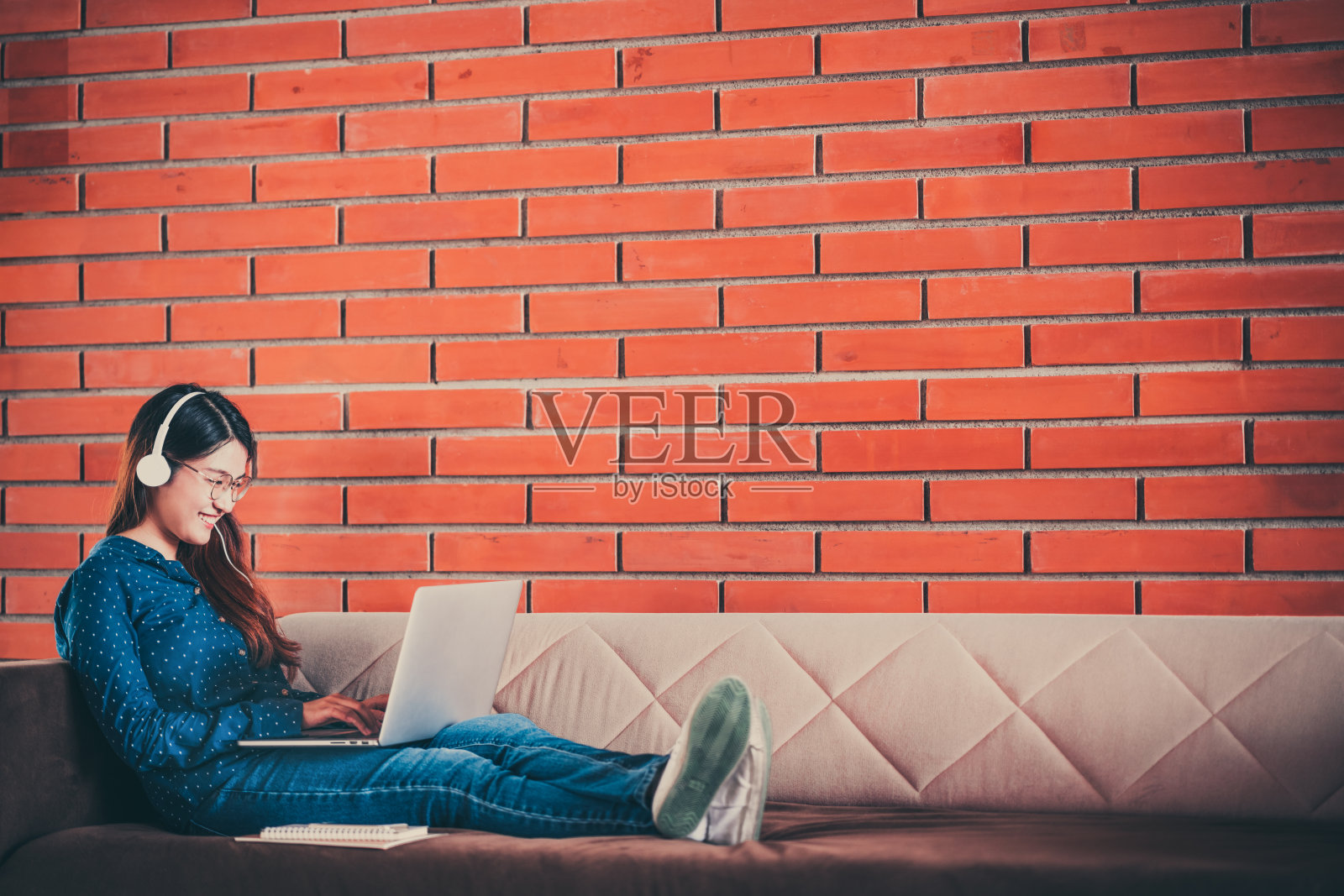 年轻的亚洲大学生女孩使用笔记本电脑，耳机，智能手机，笔记本电脑在沙发沙发上，坐在大学校园里，在砖墙上拷贝空间。教育或休闲学习生活理念照片摄影图片
