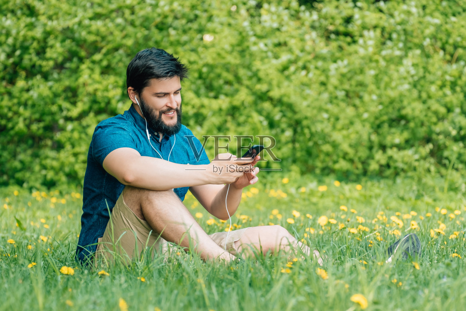 一个年轻人坐在公园里发短信。在草地上用智能手机微笑的男人照片摄影图片