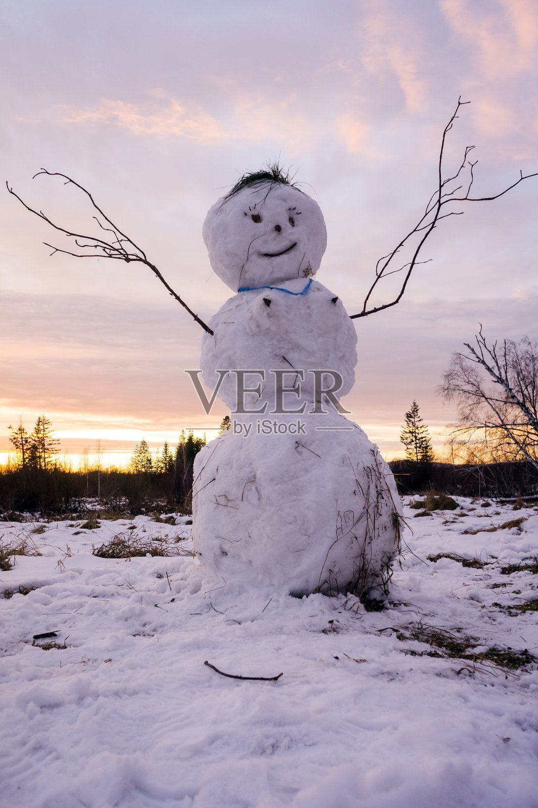 冬天的风景。夕阳西下，一个丑陋的雪人举着胳膊和树枝站在白雪覆盖的草地上。照片摄影图片