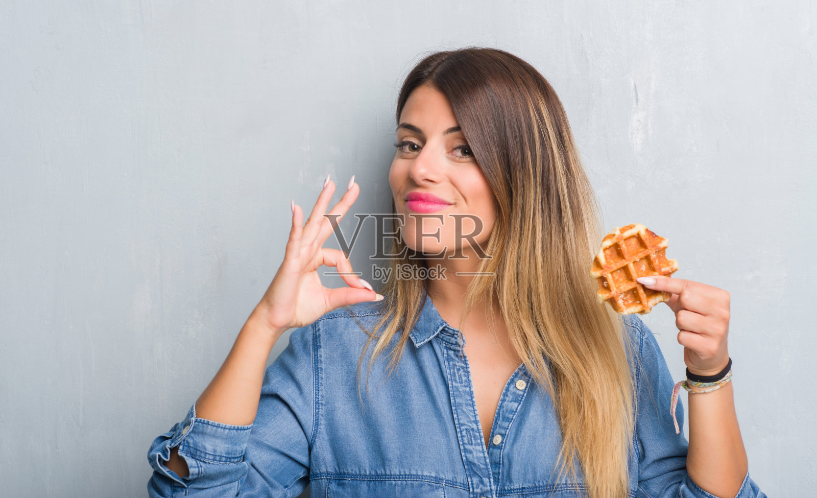 年轻的成年女子越过灰色的垃圾墙吃着比利时华夫饼，用手指做着ok的手势，很好的象征照片摄影图片