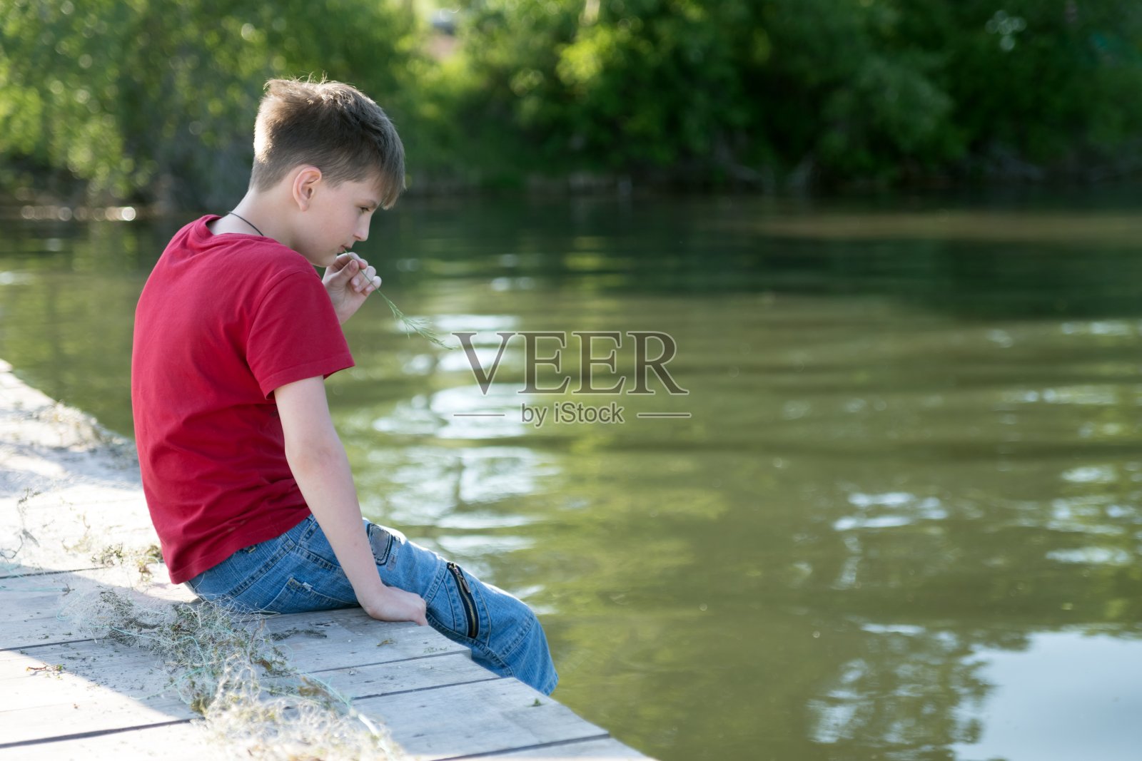 在长满树木的河岸的背景下，一个沉思的男孩坐在一个木栈桥上，把脚伸入水中，啃着一片草叶。照片摄影图片