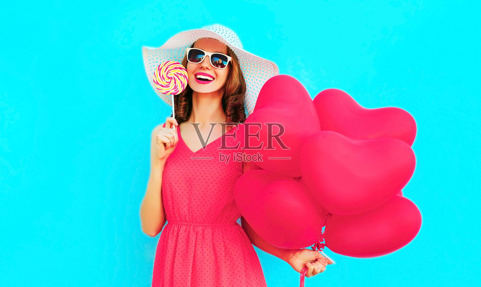 快乐微笑的年轻女子拿着棒棒糖，粉红色的心形气球在彩色的蓝色背景，戴着夏季草帽照片摄影图片