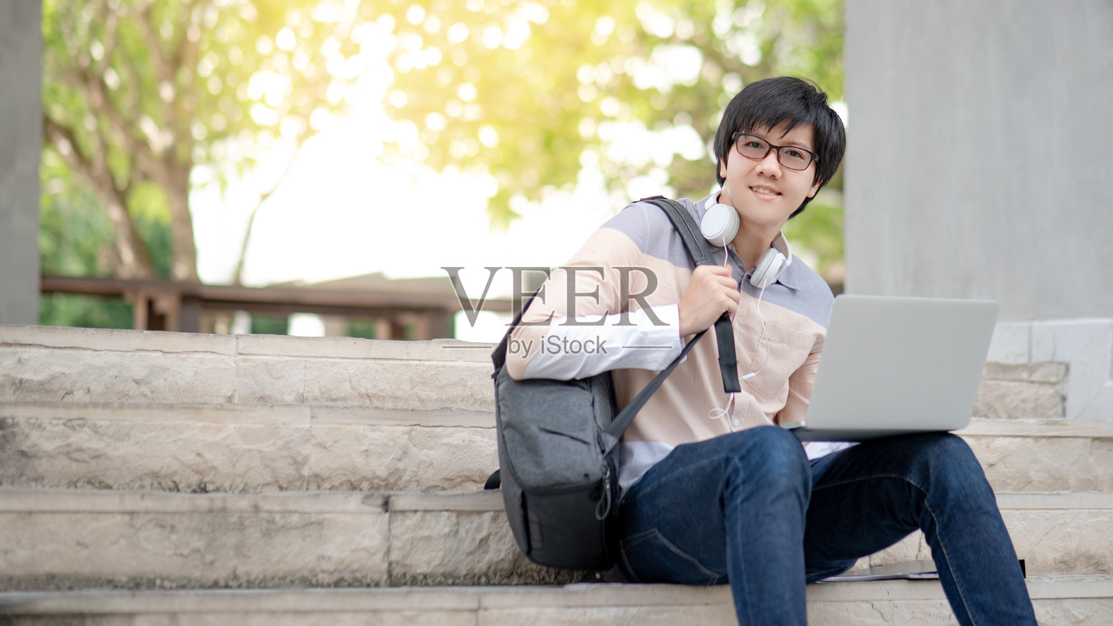 年轻的亚洲人大学学生戴着眼镜和耳机坐在楼梯上用笔记本电脑在大学大楼工作。教育机会和奖学金。学习成功的步骤。照片摄影图片