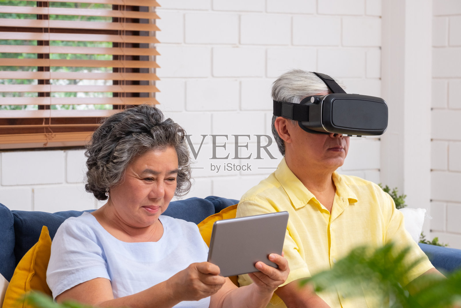 亚洲老年夫妇玩虚拟现实眼镜耳机和平板电脑看虚拟视频，在家里客厅的沙发上一起玩乐。年长技术。老在家里照片摄影图片