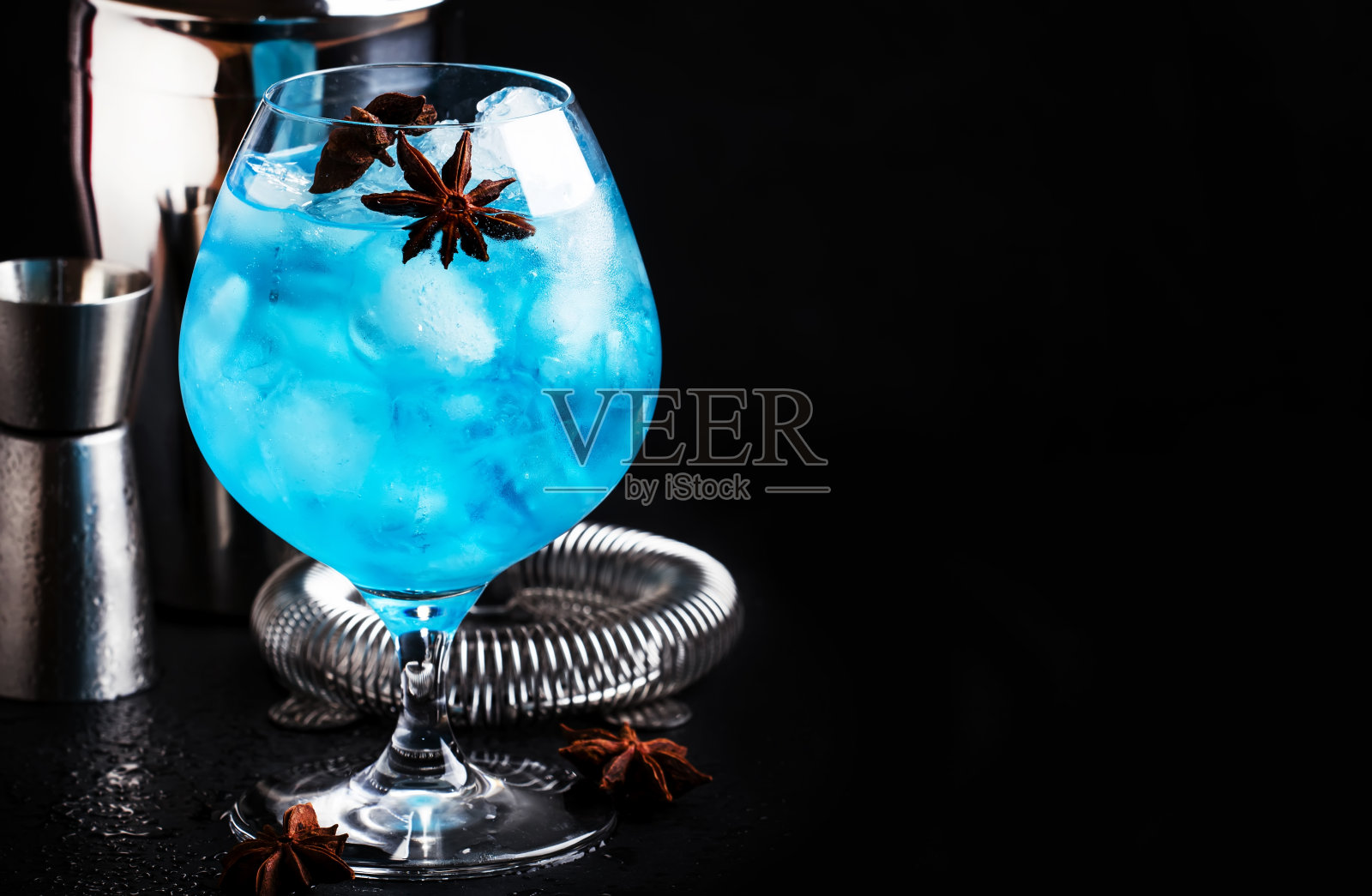 蓝色酒精鸡尾酒，有sambuca，利口酒，柠檬汁，碎冰和八角星形大葡萄酒杯与调酒师钢工具，黑色吧台背景，选择性聚焦照片摄影图片