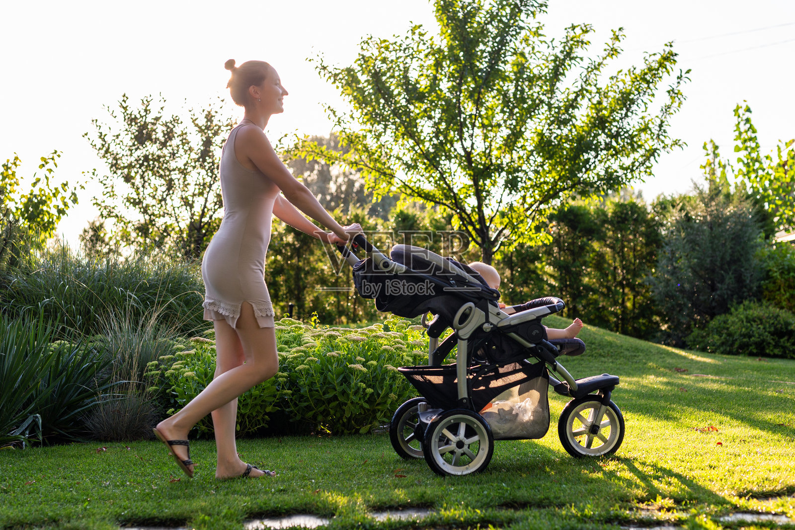 年轻漂亮的母亲与婴儿在婴儿车享受散步在绿色新鲜的花园日落。妈妈在美丽的公园里和婴儿玩得很开心。快乐的母亲概念照片摄影图片