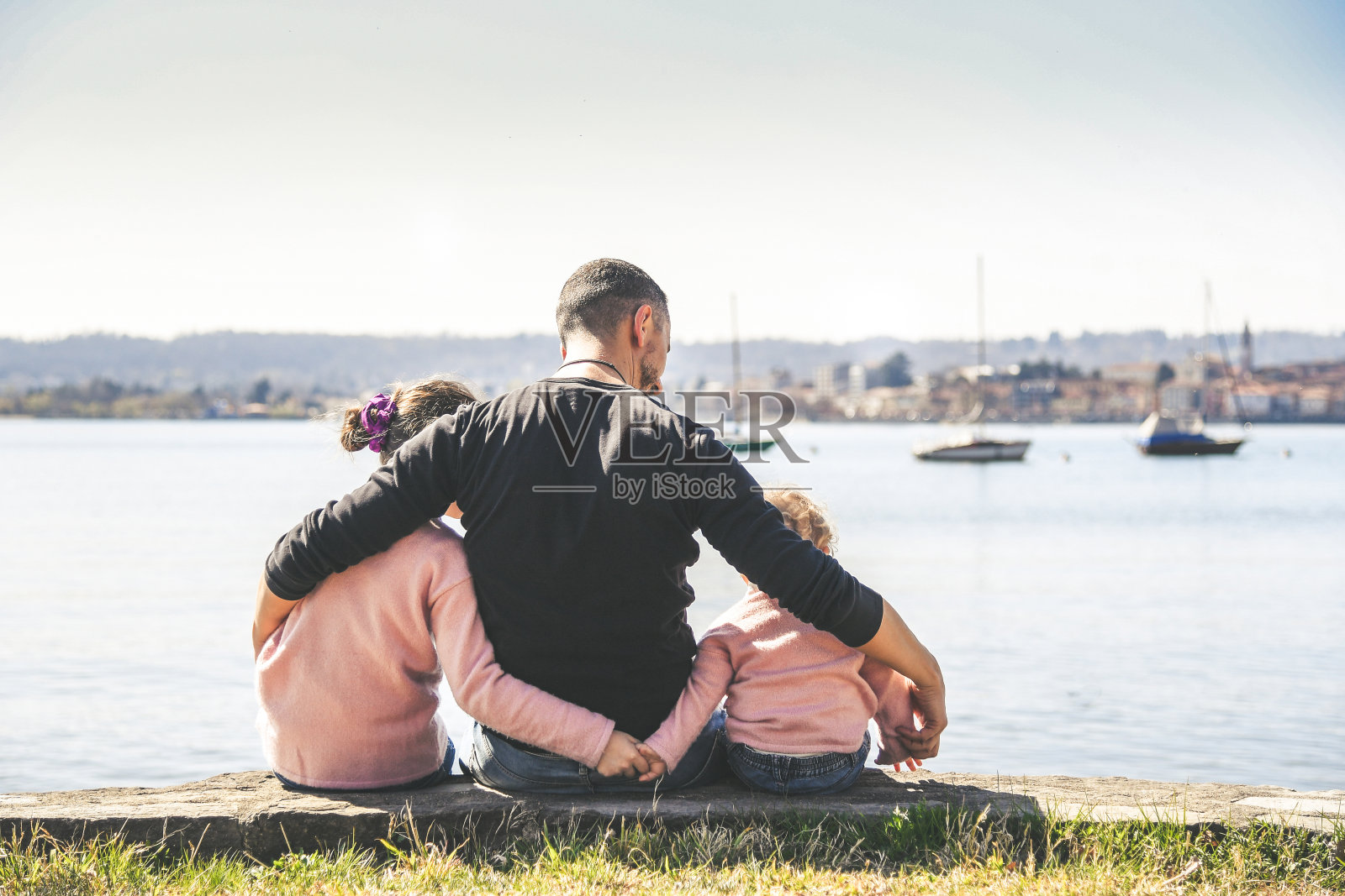 在一个阳光明媚的春日，爸爸和两个小妹妹坐在海边拥抱。父亲和儿子一起在海边。家庭爱的概念形象，关爱保护和情感结合。照片摄影图片