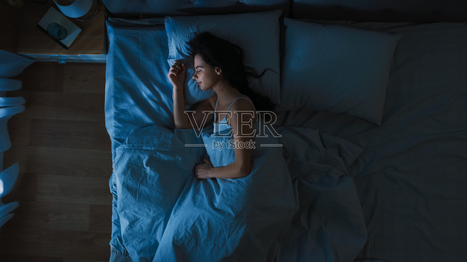 俯视图美丽的年轻女子舒适地睡在床上在他的卧室晚上。蓝色的夜间颜色和寒冷微弱的灯柱灯光透过窗户。照片摄影图片