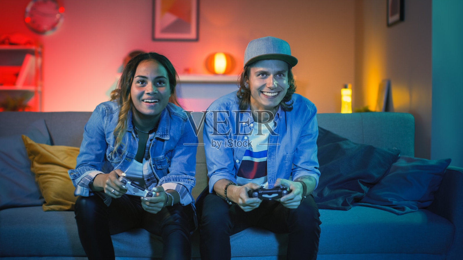 兴奋的黑人玩家女孩和年轻人坐在沙发上，在主机上玩视频游戏。他们玩无线控制器。温馨的房间，以温暖和霓虹灯照明。照片摄影图片