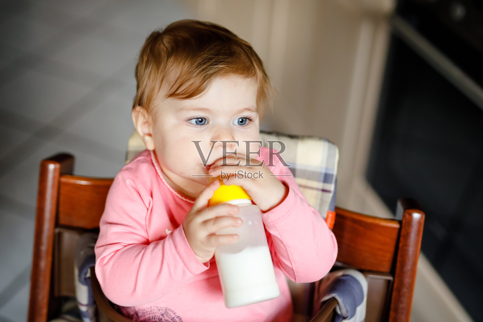 可爱可爱的小女孩拿着奶瓶喝配方奶。首先是给婴儿的食物。新生儿，坐在家庭厨房的椅子上。健康婴儿和奶瓶喂养的概念照片摄影图片