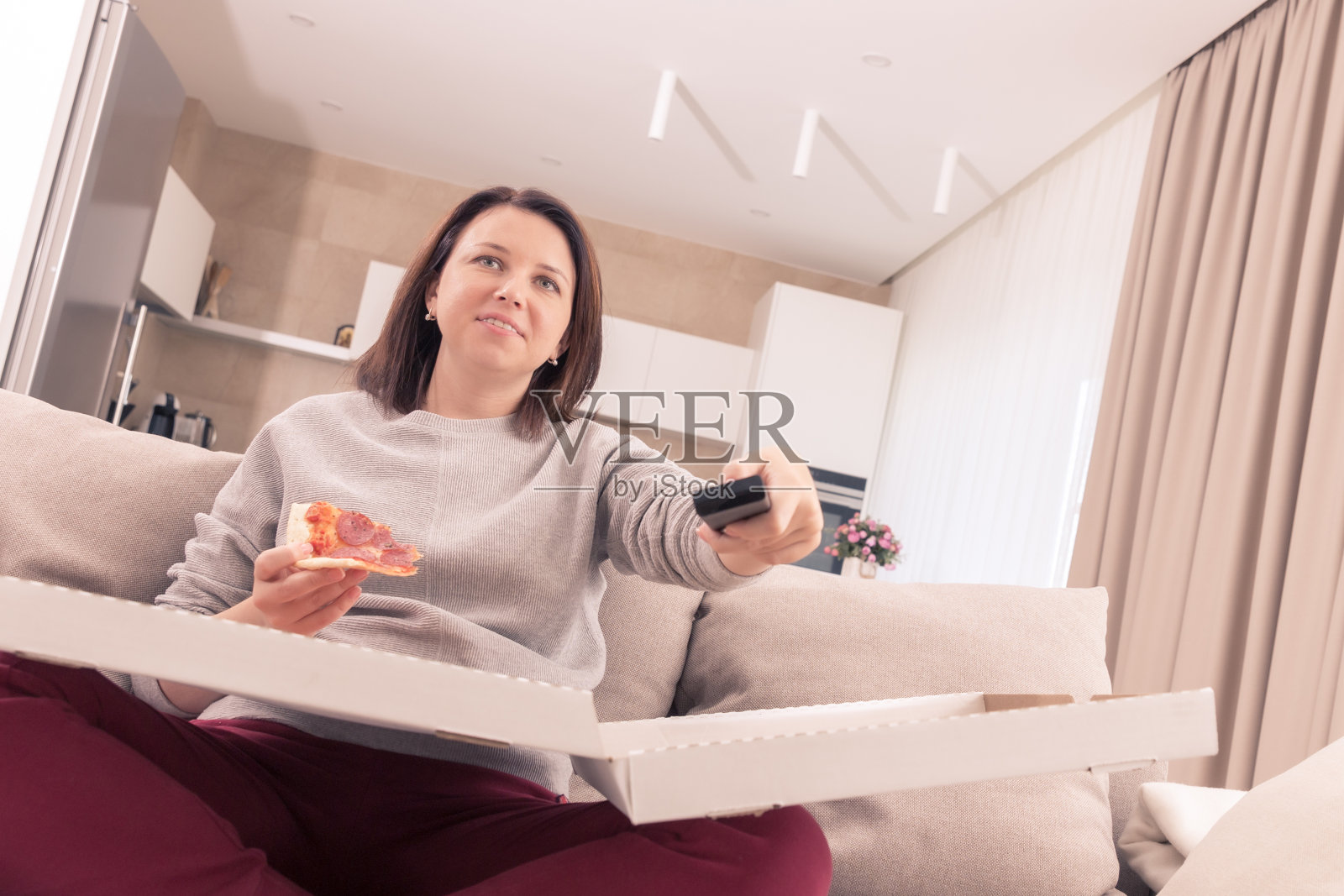 漂亮女人在家里一边吃披萨一边看电视一边拿着遥控器，橙色调照片摄影图片