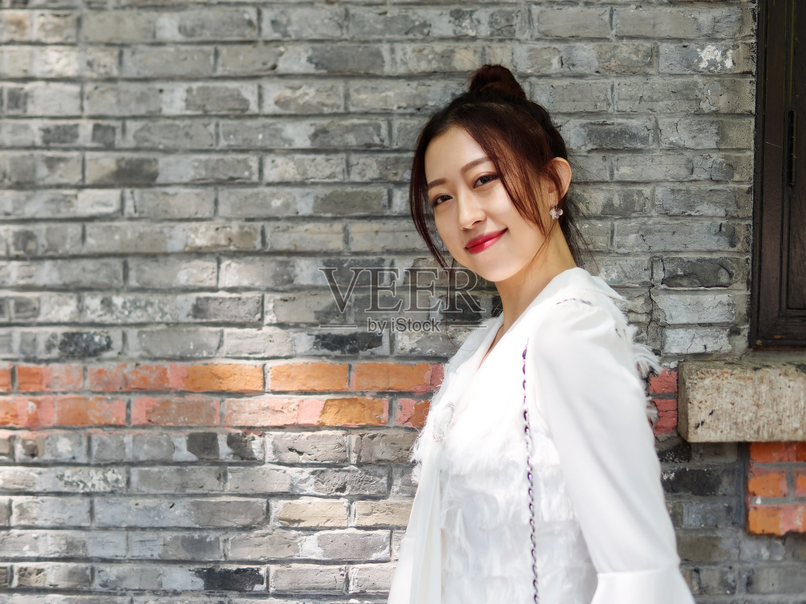 在阳光明媚的日子里，美丽的中国年轻女子穿着白色的衣服对着镜头微笑，背景是砖墙。户外时尚写真魅力时尚女士。照片摄影图片