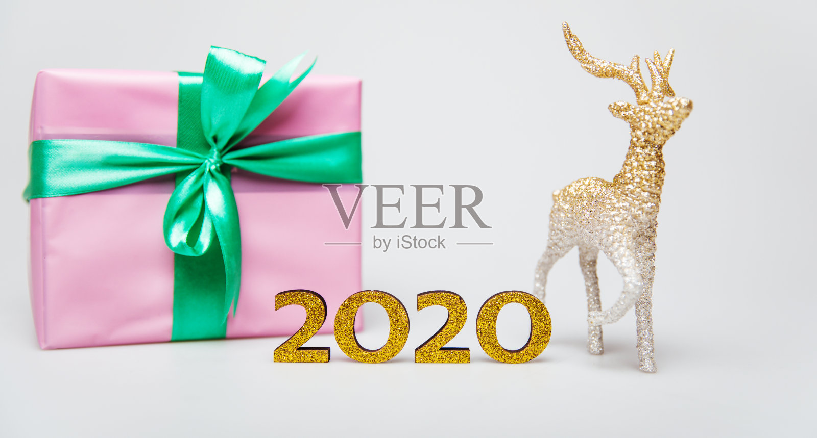 每年的这个时候。一个用粉色包装纸包裹的绿色丝带，一个金光闪闪的金色小鹿雕像和2020年的金色数字组成。照片摄影图片