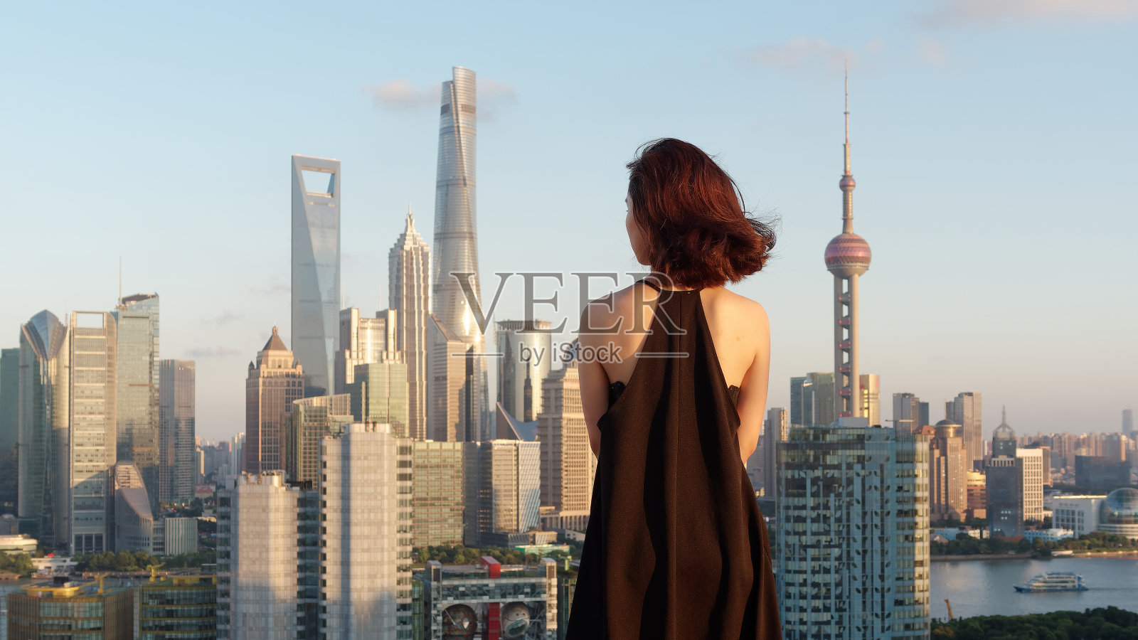 后视图美丽的年轻女子在美丽的黑色连衣裙摆姿势在大厦屋顶与模糊的上海外滩地标建筑的背景。情感、人、美、生活理念。照片摄影图片