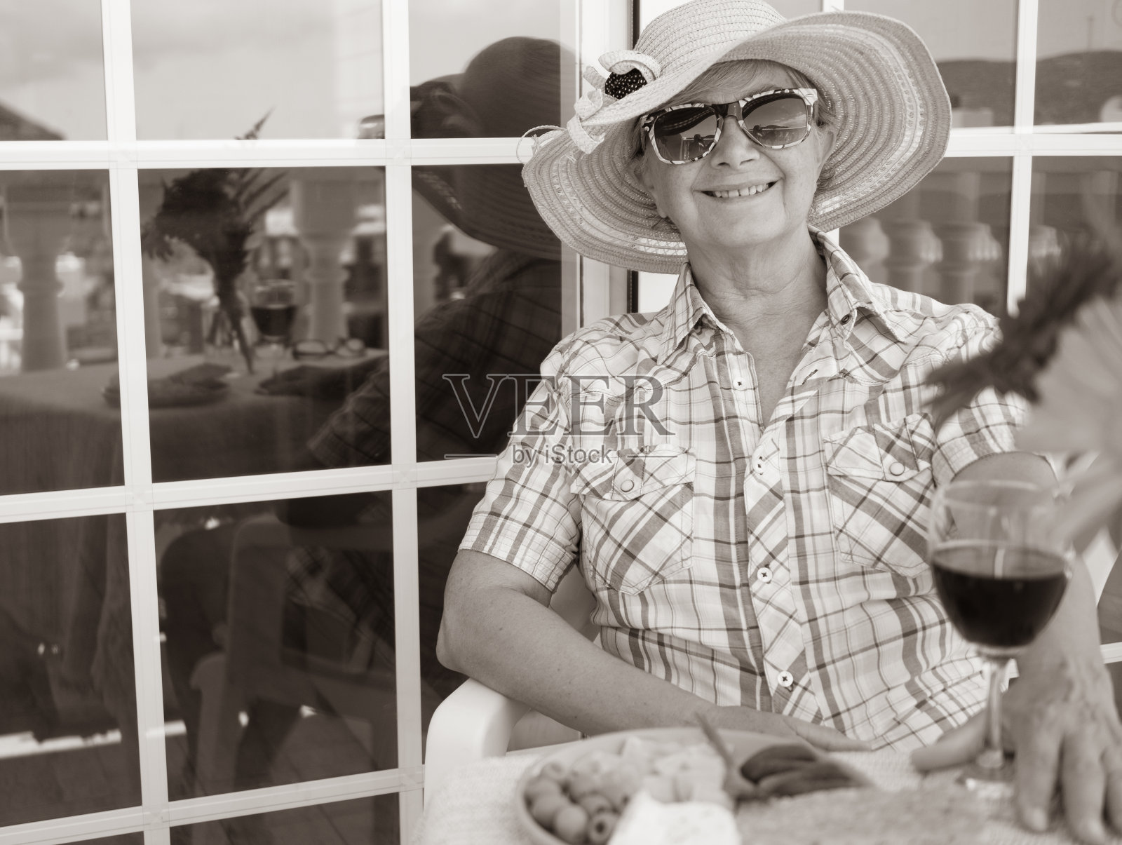 成年老奶奶在露天的露台上吃饭。手里拿着红酒杯，享受着一些开胃菜。粉红色的夏日帽子下的微笑照片摄影图片