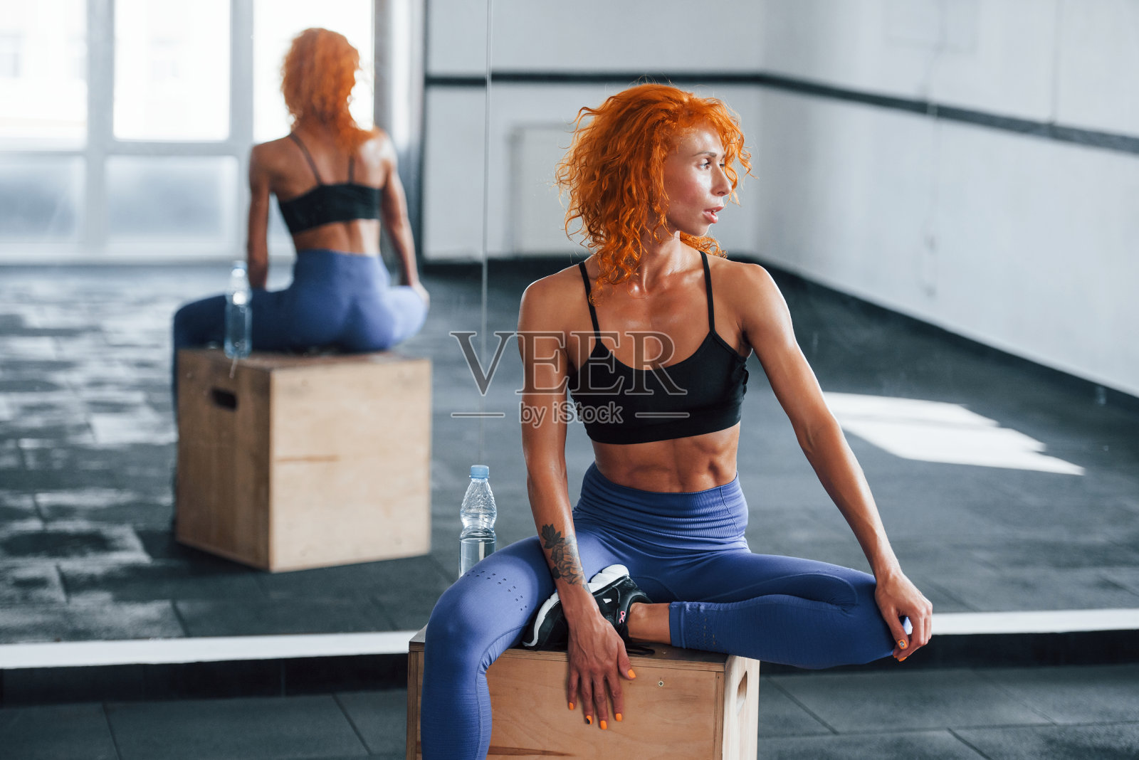坐下来休息一下，旁边放一瓶水。喜欢运动的红发女孩白天去健身房健身。肌肉发达的身体类型照片摄影图片