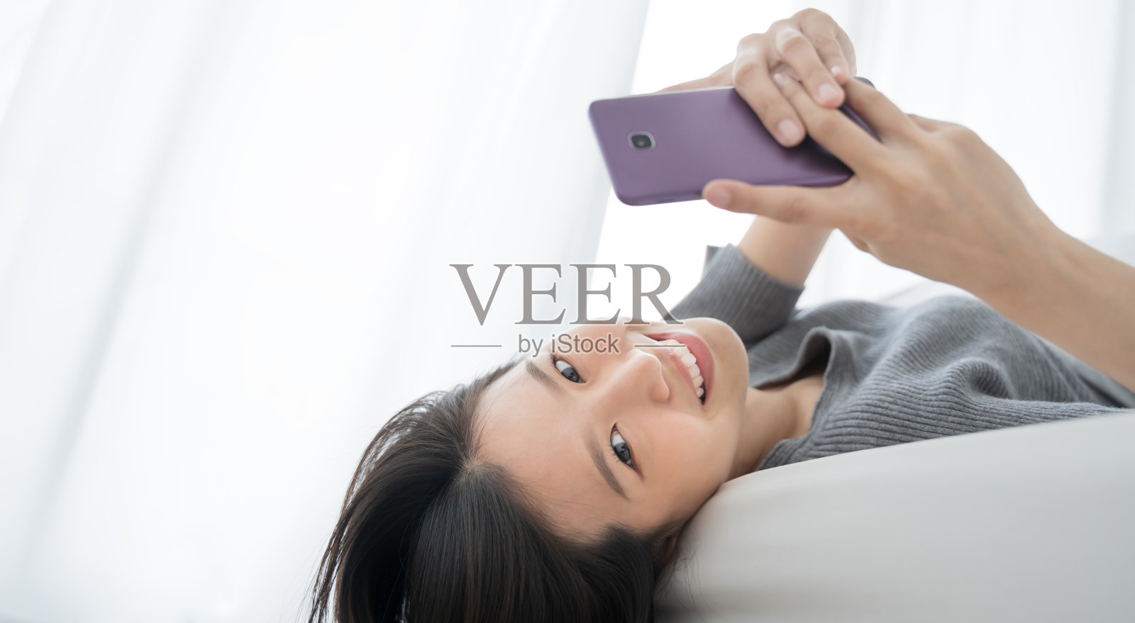 年轻的亚洲女人使用智能手机在卧室快乐和微笑。照片摄影图片