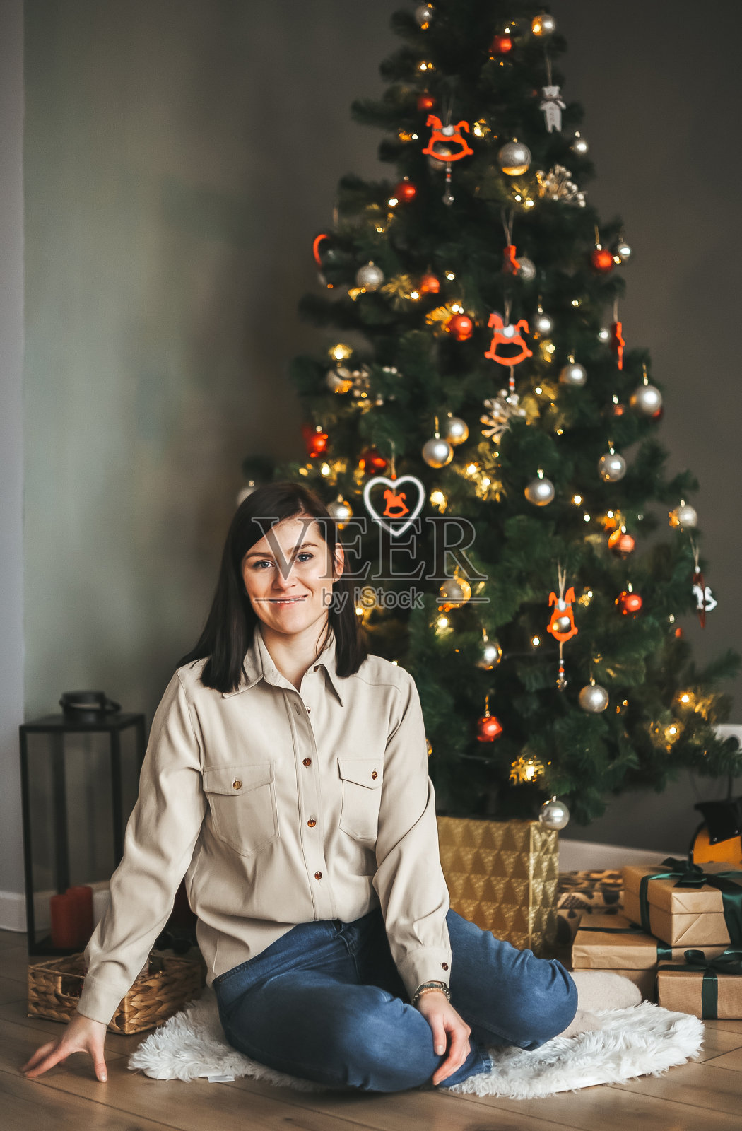 美丽的女人正坐在圣诞树旁的白色小地毯上。照片摄影图片