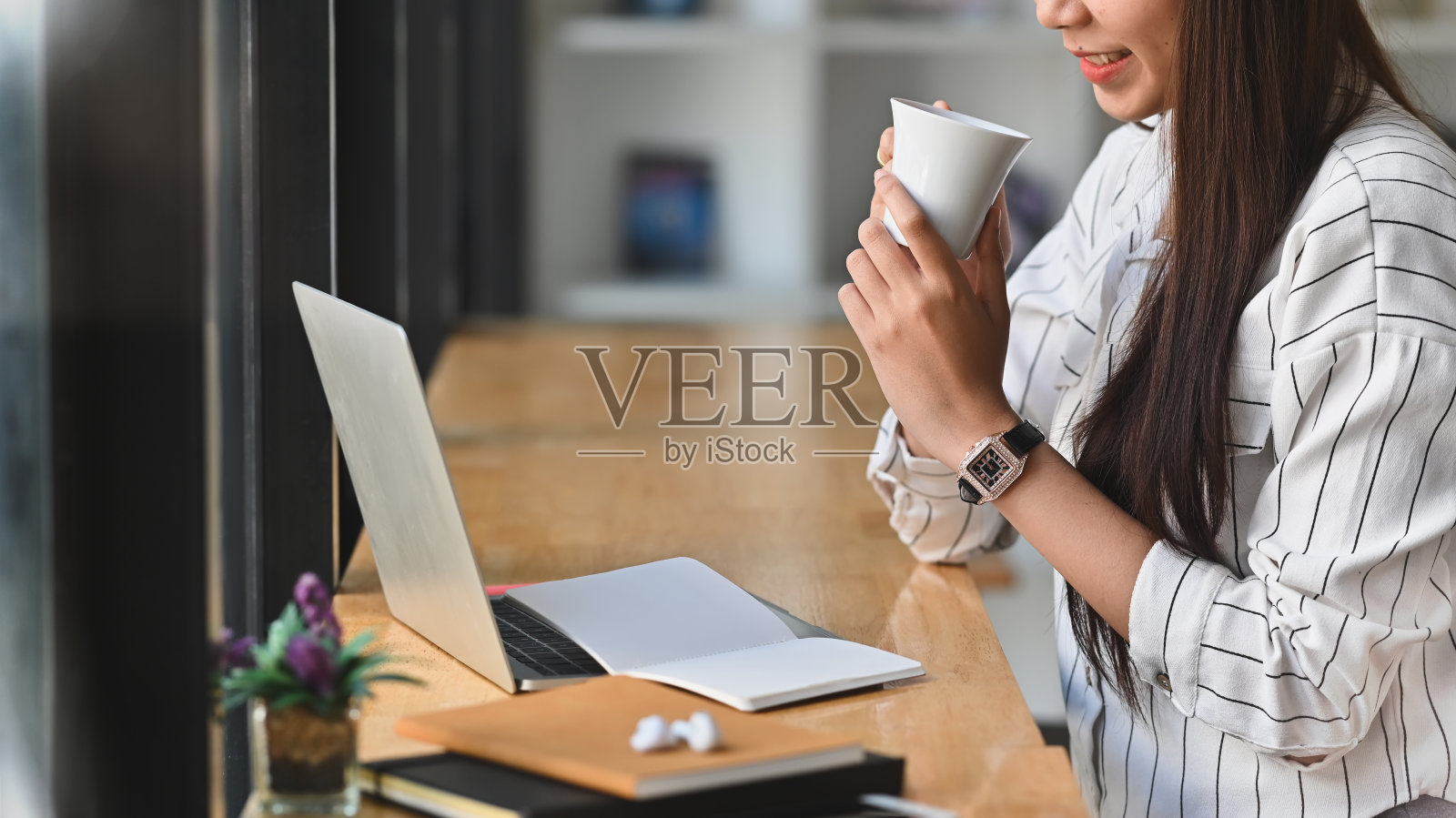 侧拍的年轻的商业女性，而拿着一个咖啡杯，看着笔记本电脑在现代的办公室/咖啡馆。照片摄影图片