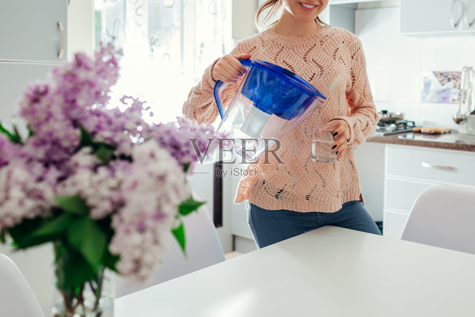 在厨房里，一个妇女把过滤水壶里的水倒进玻璃杯里。现代厨房设计。健康的生活方式照片摄影图片
