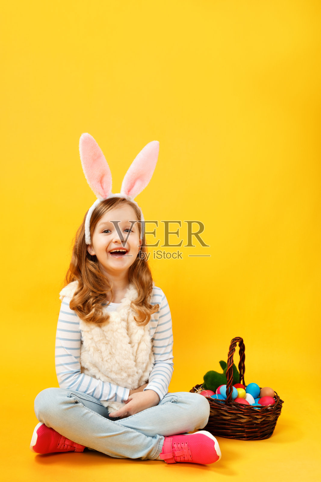 快乐的小女孩拿着一篮子复活节彩蛋坐在地板上。快乐可爱美丽的小兔子长在耳朵上的黄色背景画室里照片摄影图片