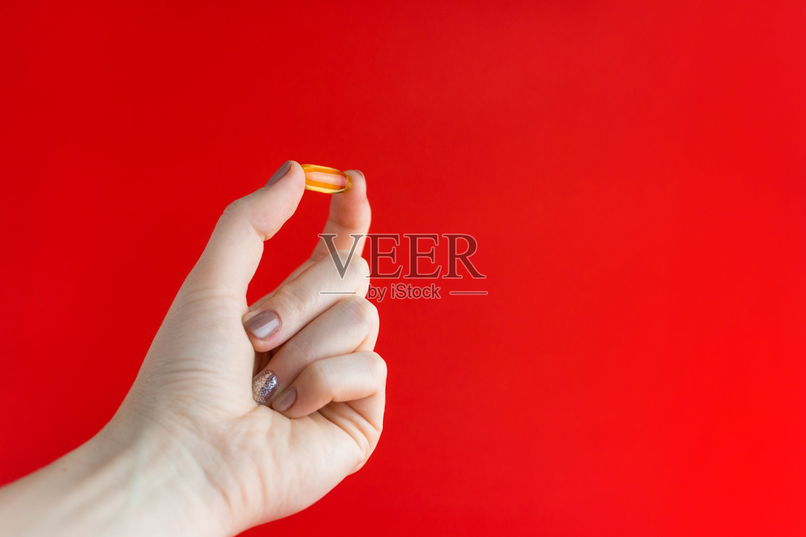 黄色药丸夹在两指间。一个女孩的手拿着一颗透明的药丸在红色的背景。金色鱼油胶囊，女性手掌中的欧米伽-3鱼油照片摄影图片