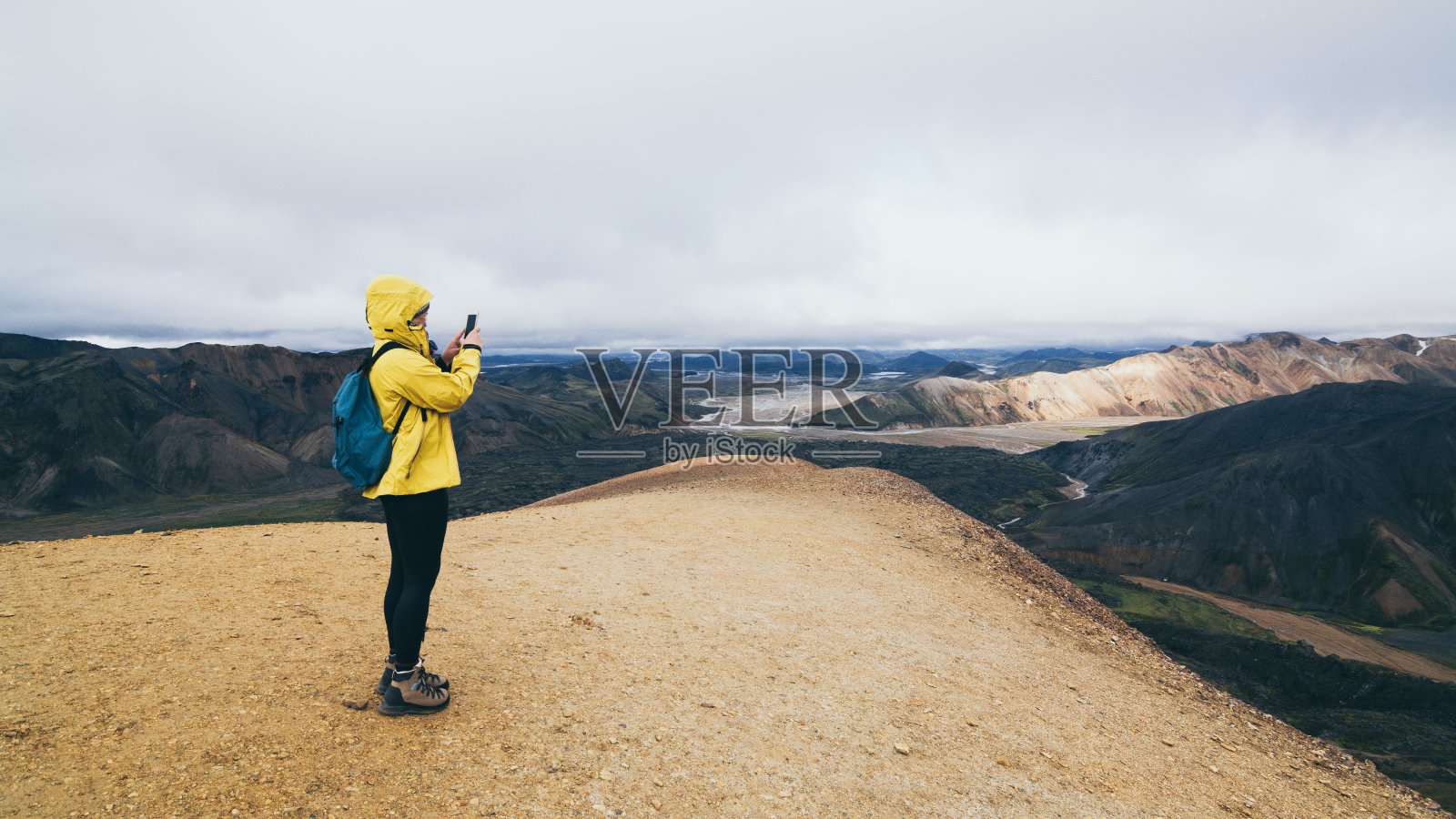 一名身穿黄色雨衣的妇女徒步旅行并在冰岛的Landmannalaugar国家公园里拍摄五彩缤纷的山脉照片摄影图片