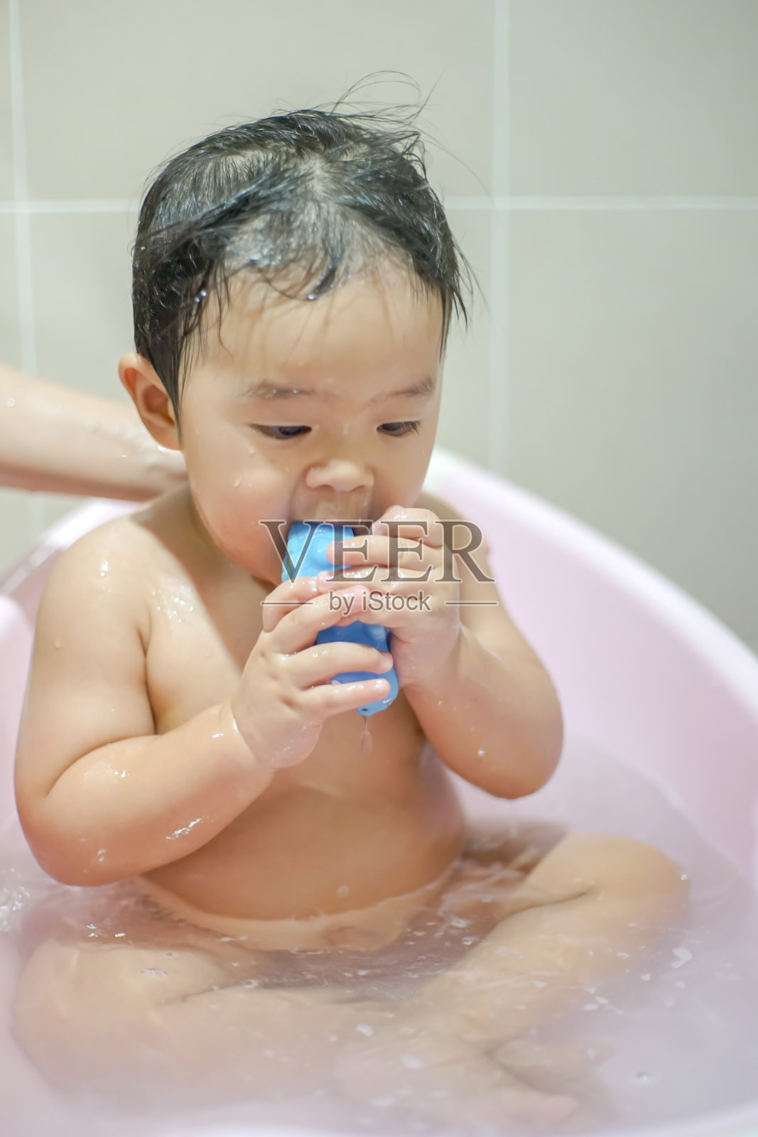 一个小男孩正在洗澡，在粉红色的浴缸里咬玩具。照片摄影图片