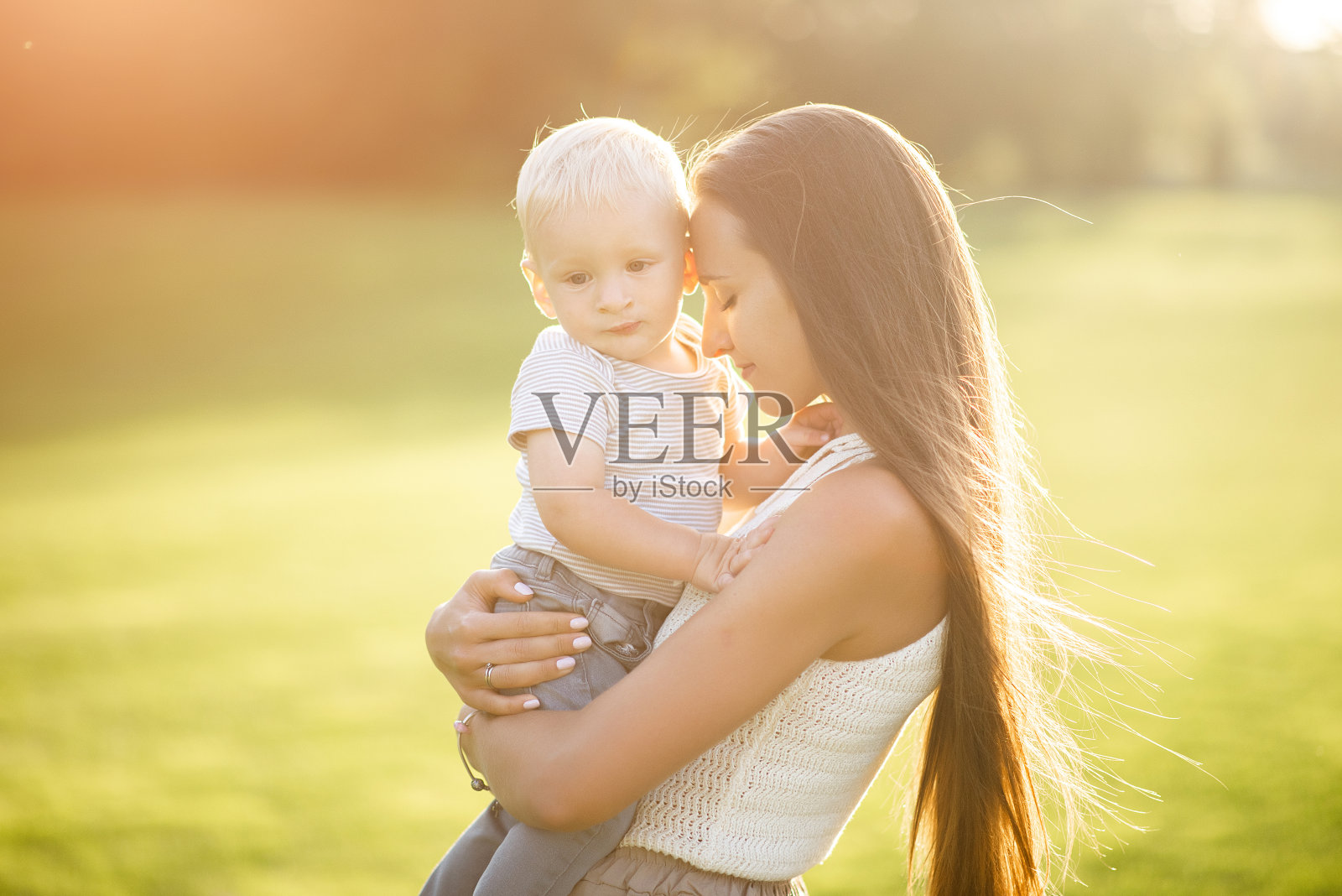 一位长发的母亲轻轻地把她的小儿子抱在怀里。在自然光线下的公园里拍照照片摄影图片