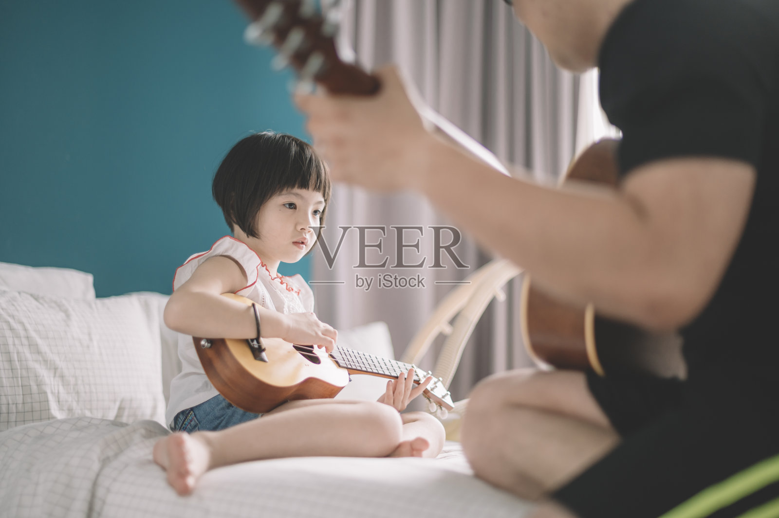 周末，在卧室的床上，一个亚洲华人的小女儿在向她的父亲学习弹奏尤克里里琴照片摄影图片