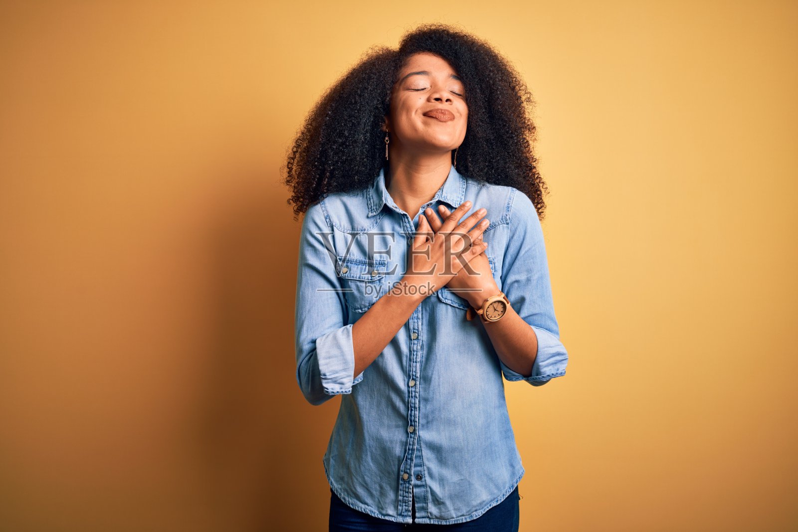 年轻美丽的非洲裔美国女人与非洲式的头发站在黄色孤立的背景微笑与手在胸前闭上眼睛和感激的手势在脸上。健康的概念。照片摄影图片