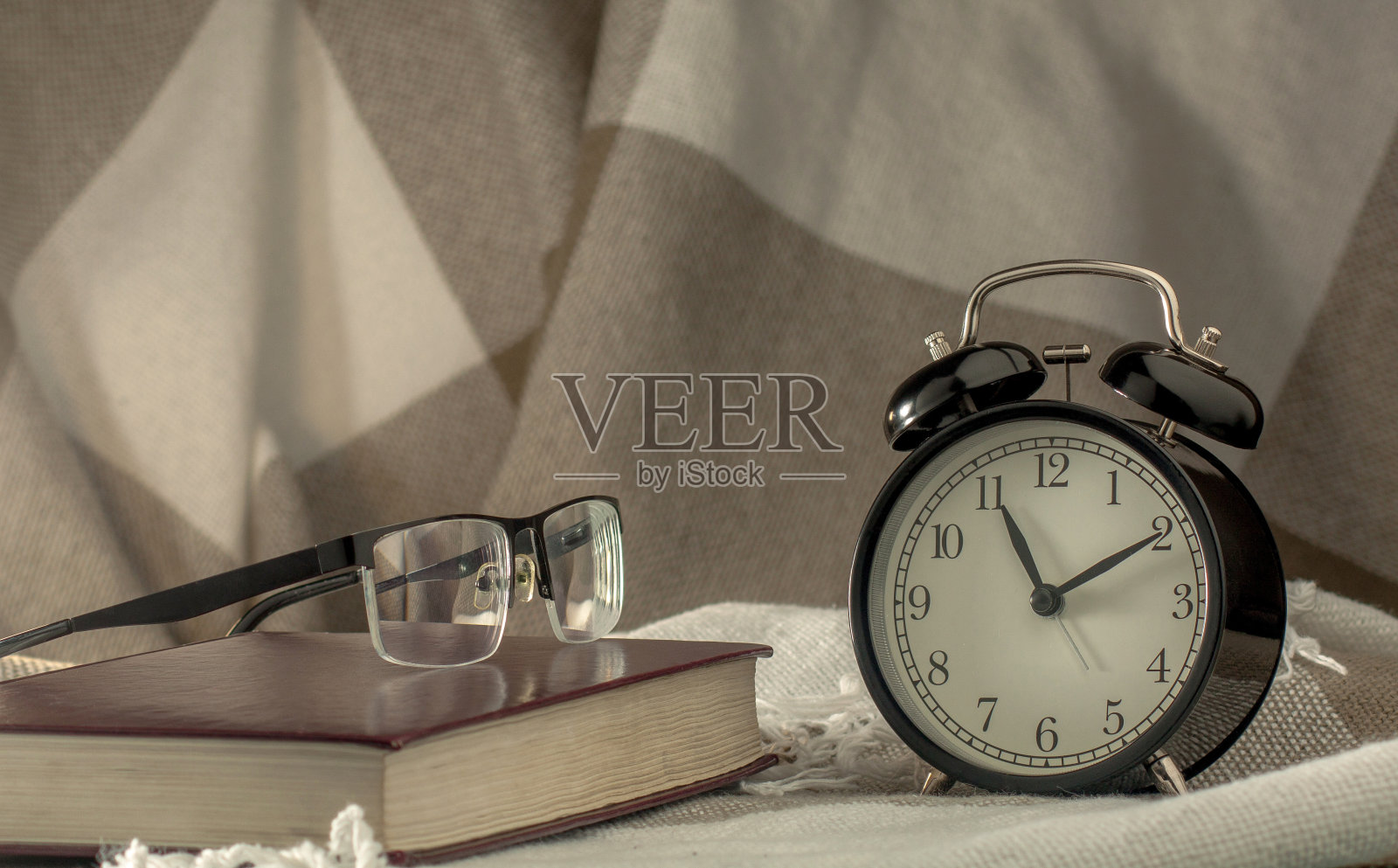 一个黑色的老式闹钟，上面放着一本棕色的大书和一副眼镜，背景是一件格子花呢照片摄影图片