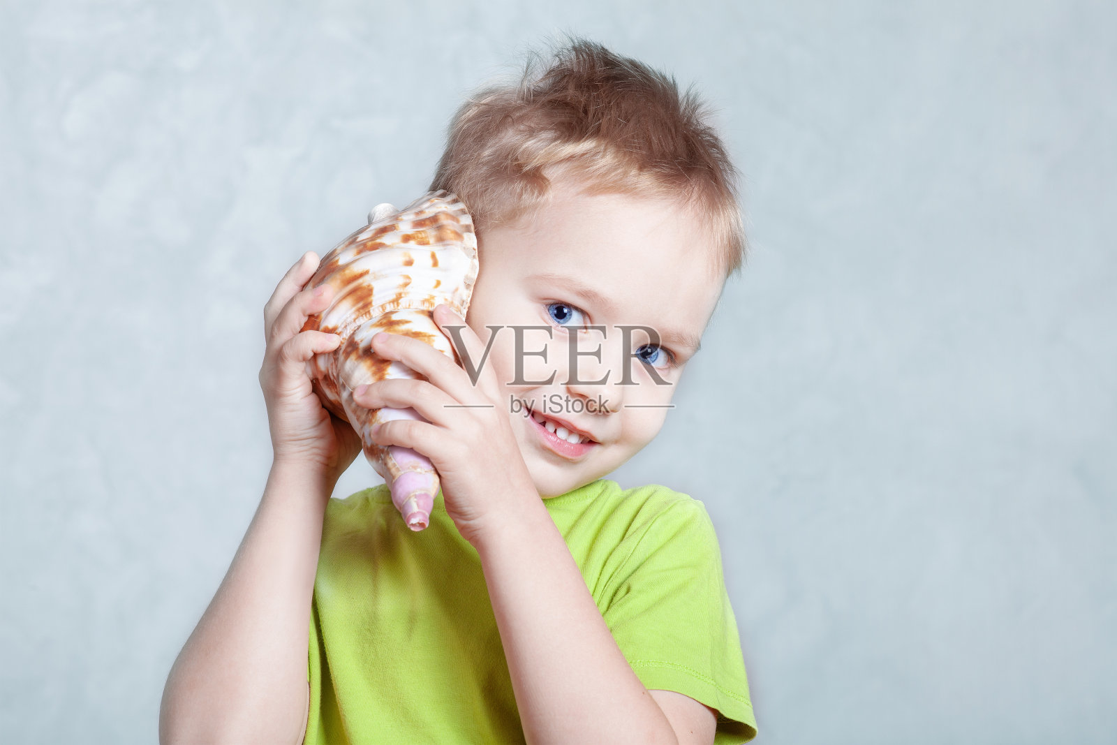 这个金发碧眼的可爱的白人小男孩一边听着贝壳的声音，一边微笑着。照片摄影图片