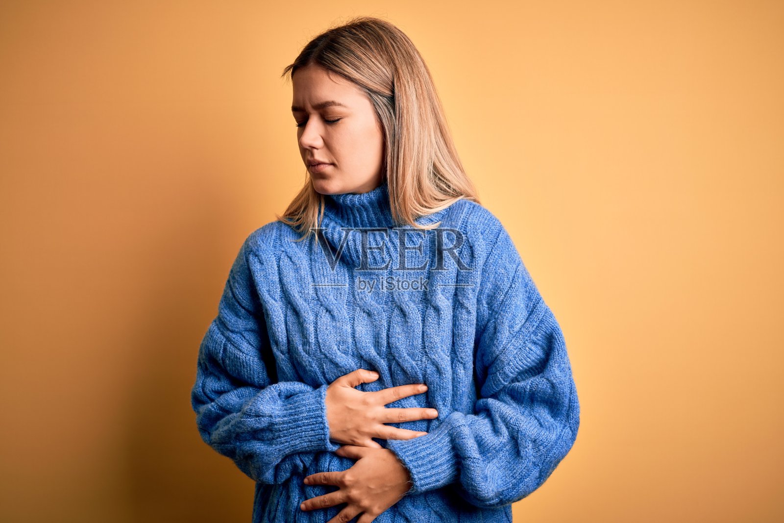 年轻美丽的金发女人穿着高领毛衣在黄色孤立的背景与手对胃，因为消化不良，痛苦的疾病感觉不舒服。疼痛的概念。照片摄影图片