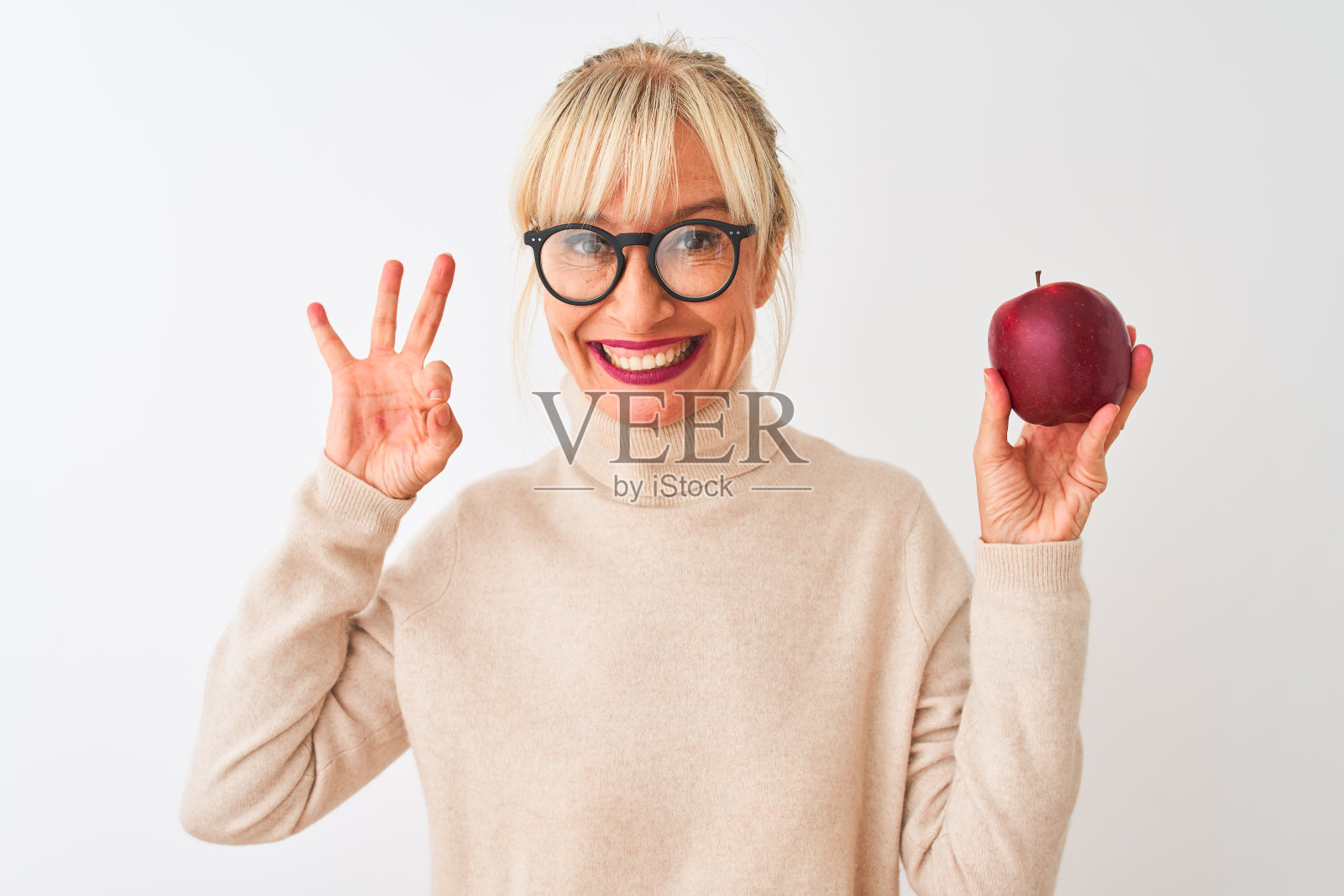 中年妇女戴着眼镜，拿着苹果站在孤立的白色背景下用手指做ok手势，很好的象征照片摄影图片
