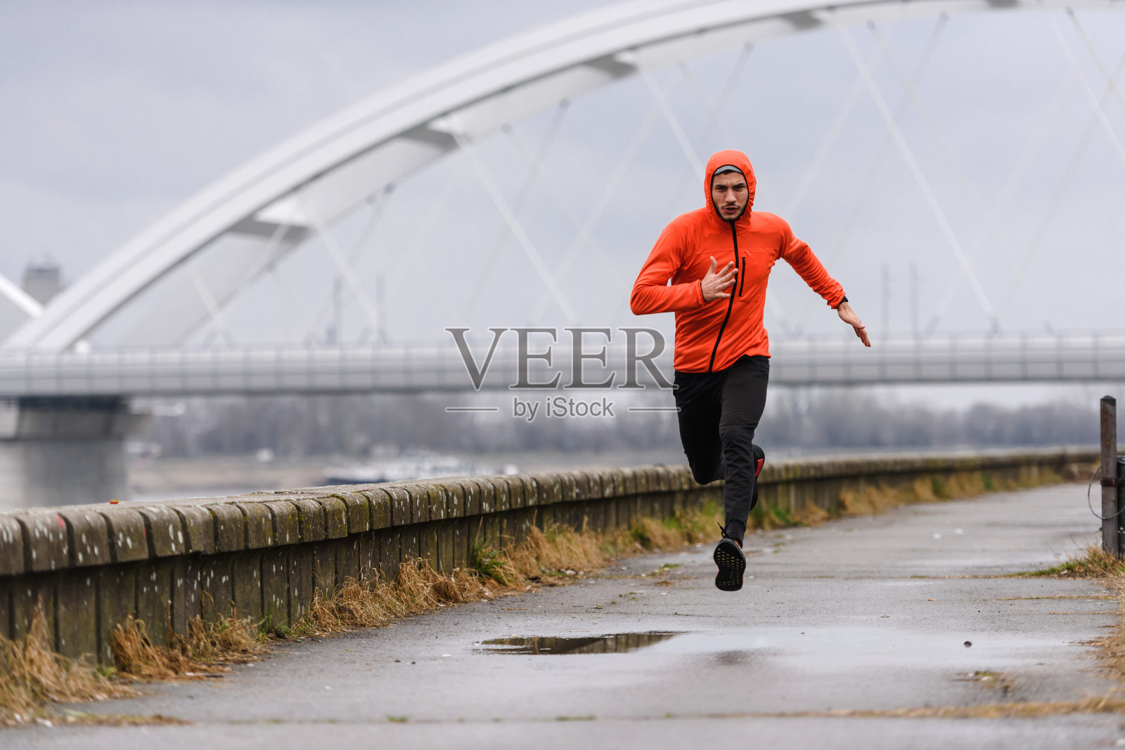 英俊的年轻运动员穿着橙色的风衣沿着河跑得很快照片摄影图片